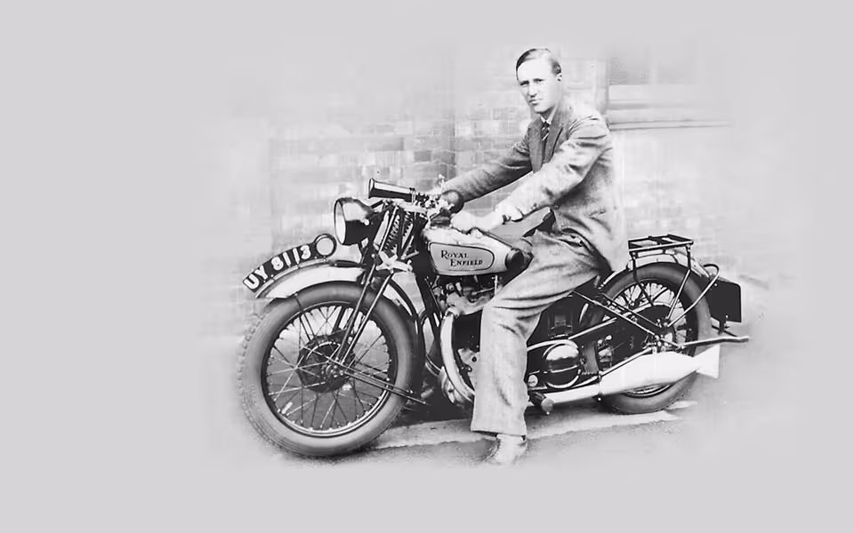 Černobílá historická fotografie muže v obleku sedícího na motocyklu Royal Enfield před cihlovou zdí.