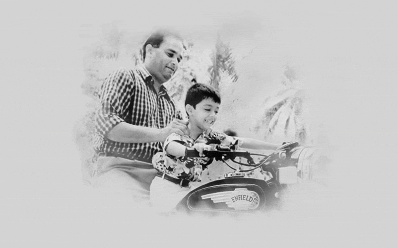 Černobílý snímek muže v kostkované košili, který učí chlapce jezdit na motocyklu Royal Enfield v přírodním prostředí.
