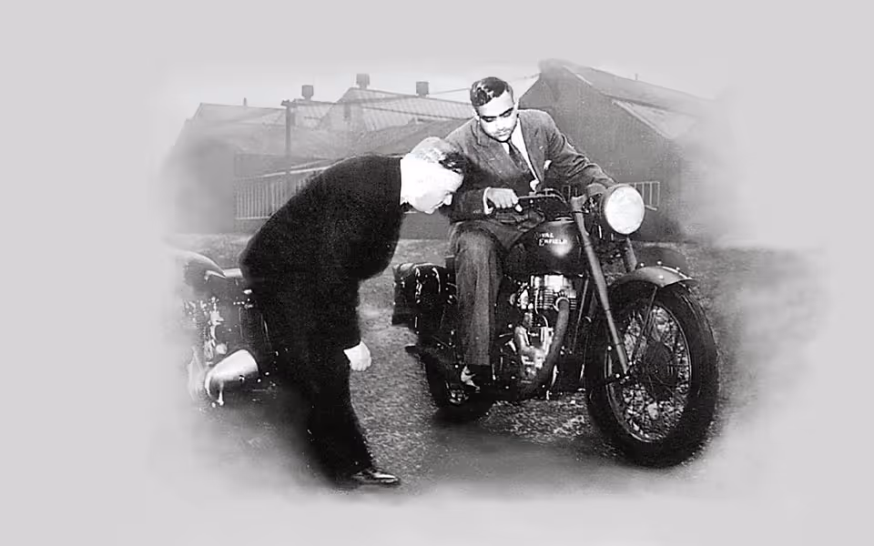 Černobílá historická fotografie muže v obleku sedícího na motocyklu Royal Enfield, zatímco druhý muž v tmavém oblečení se sklání a pozoruje přední kolo motocyklu.