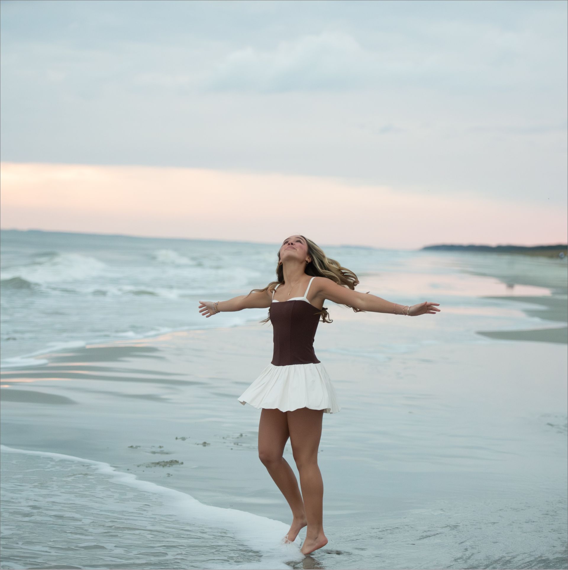 Woman in a brown and white dress standing barefoot on the beach with arms outstretched looking up at the sky during sunset.