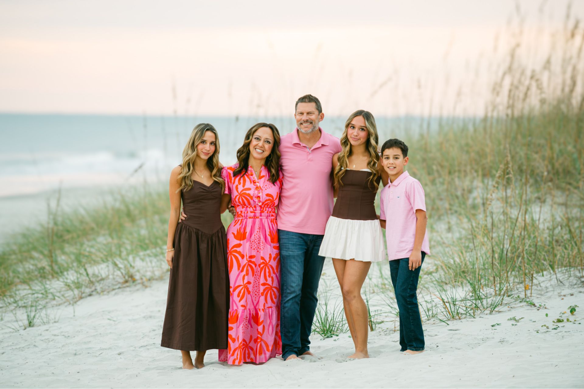 Family of five standing barefoot on a sandy beach with ocean and grass dunes in background.