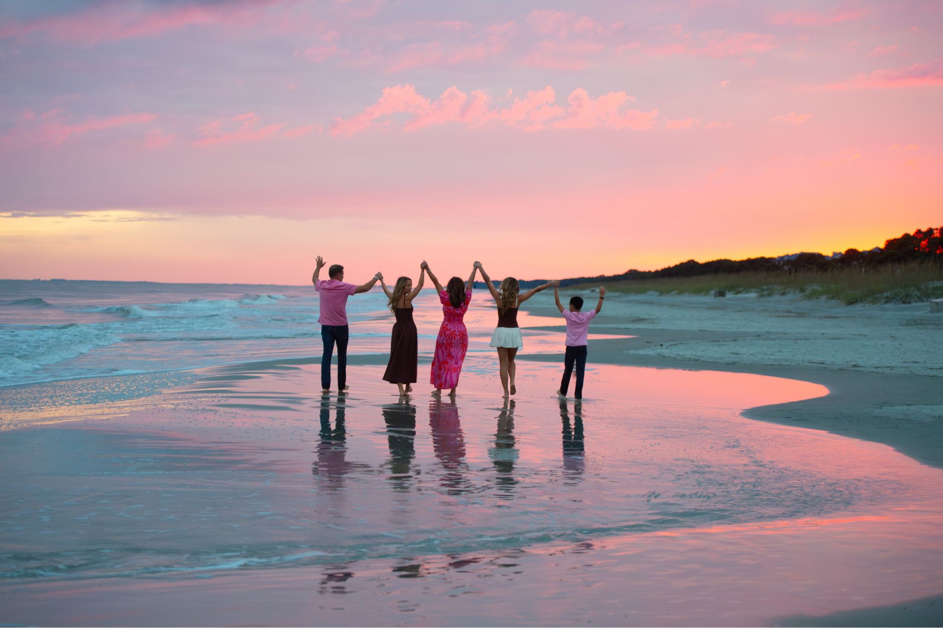 Five people holding hands and raising arms while walking on a beach at colorful sunset.
