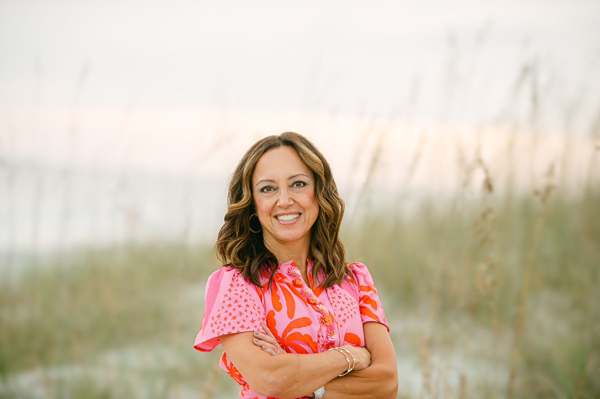 Smiling woman with wavy brown hair in a pink and orange patterned dress standing with crossed arms outdoors.