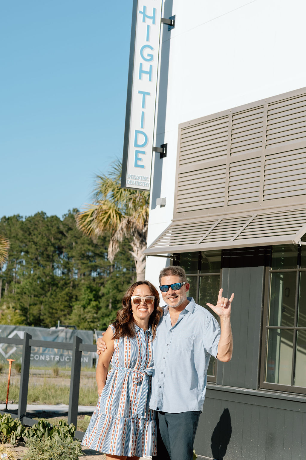 Smiling man and woman wearing sunglasses standing outside a building with a High Tide Pediatric Dentistry sign.