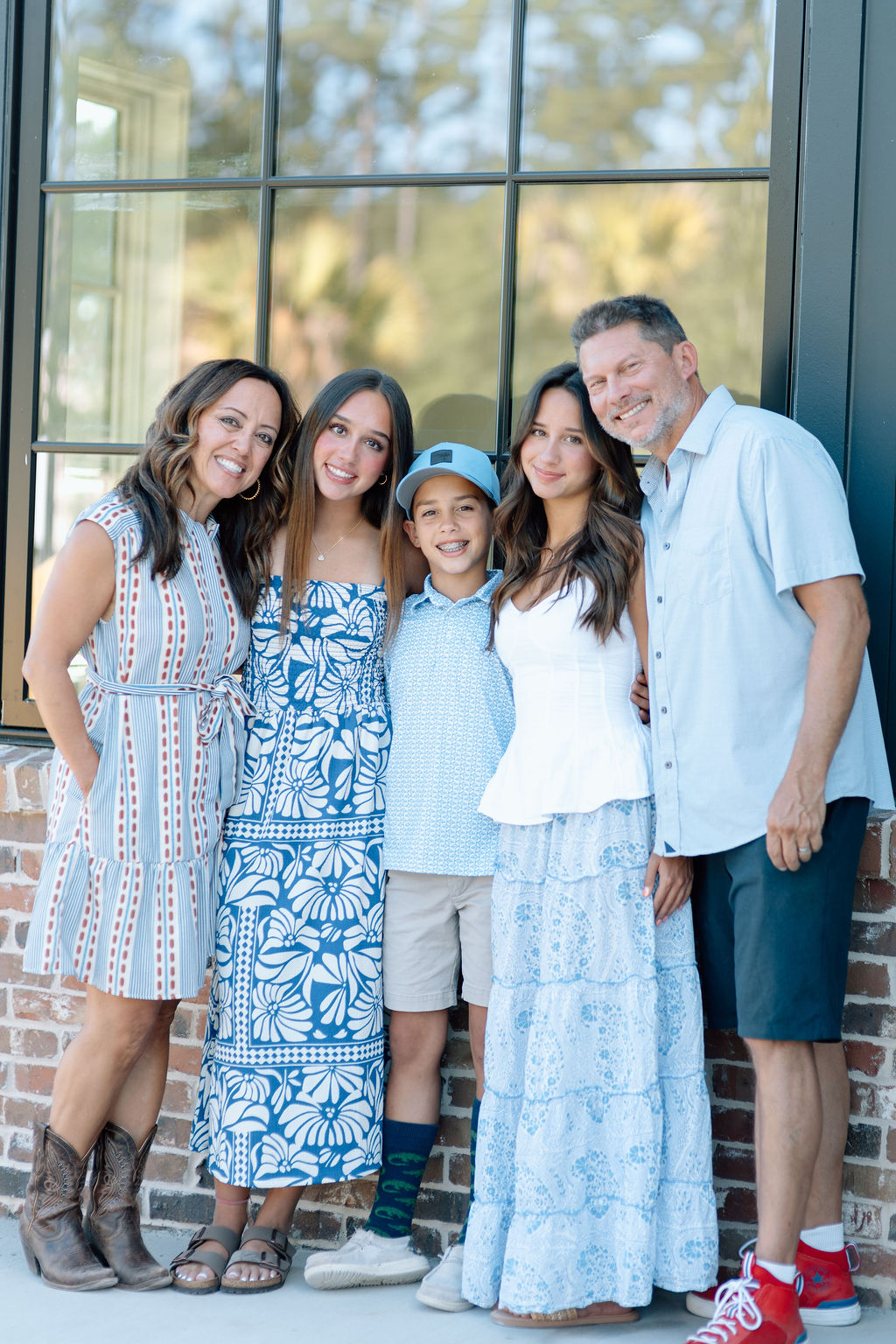 Smiling family of five posing outdoors in front of large window and brick wall.