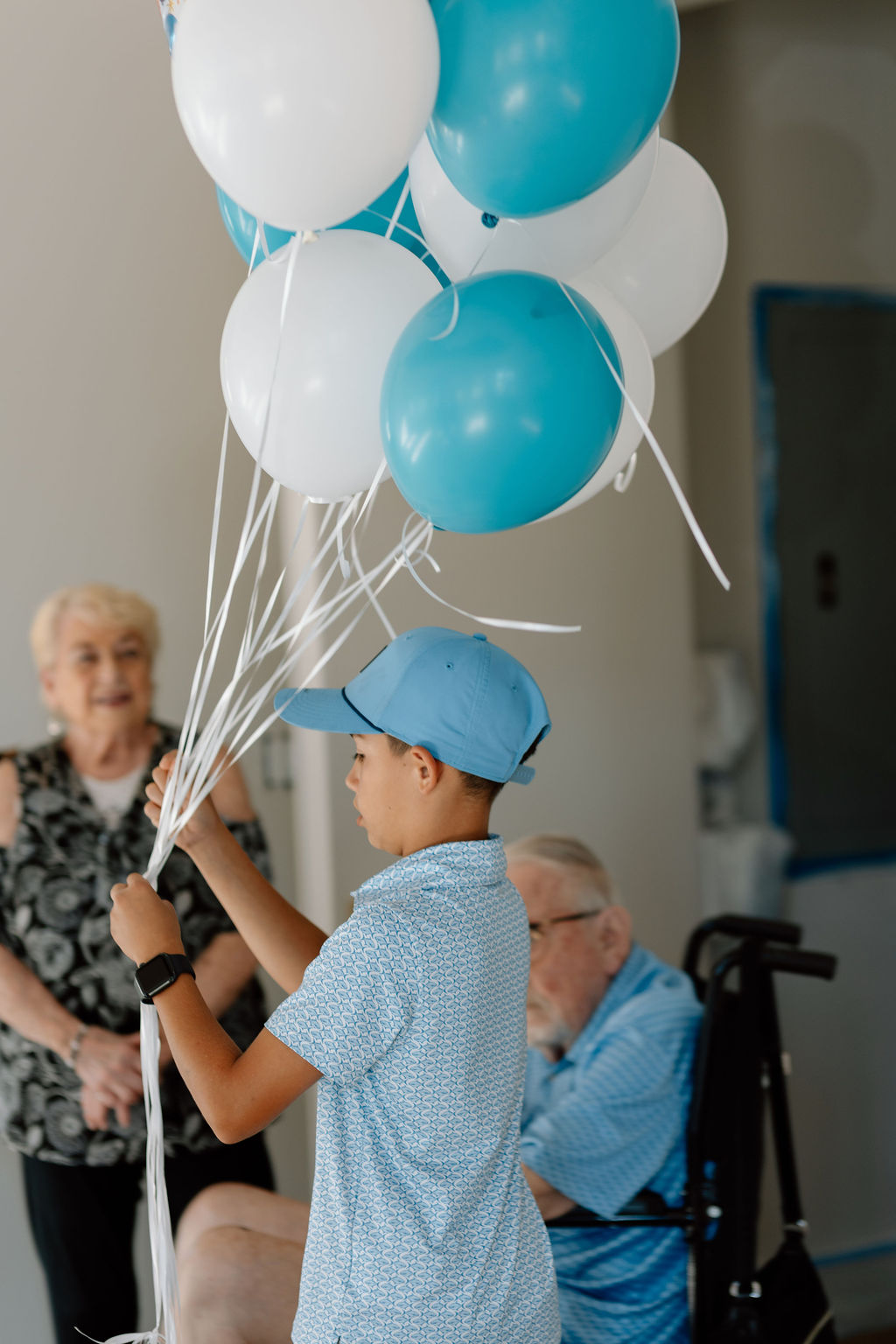 Boy in blue shirt and cap holding white and blue balloons near elderly people indoors.
