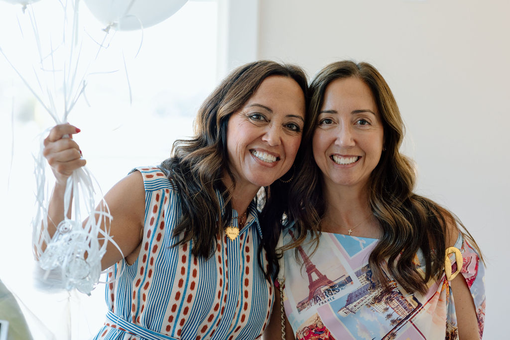 Two smiling women standing close, one holding white balloons, both wearing colorful patterned dresses.