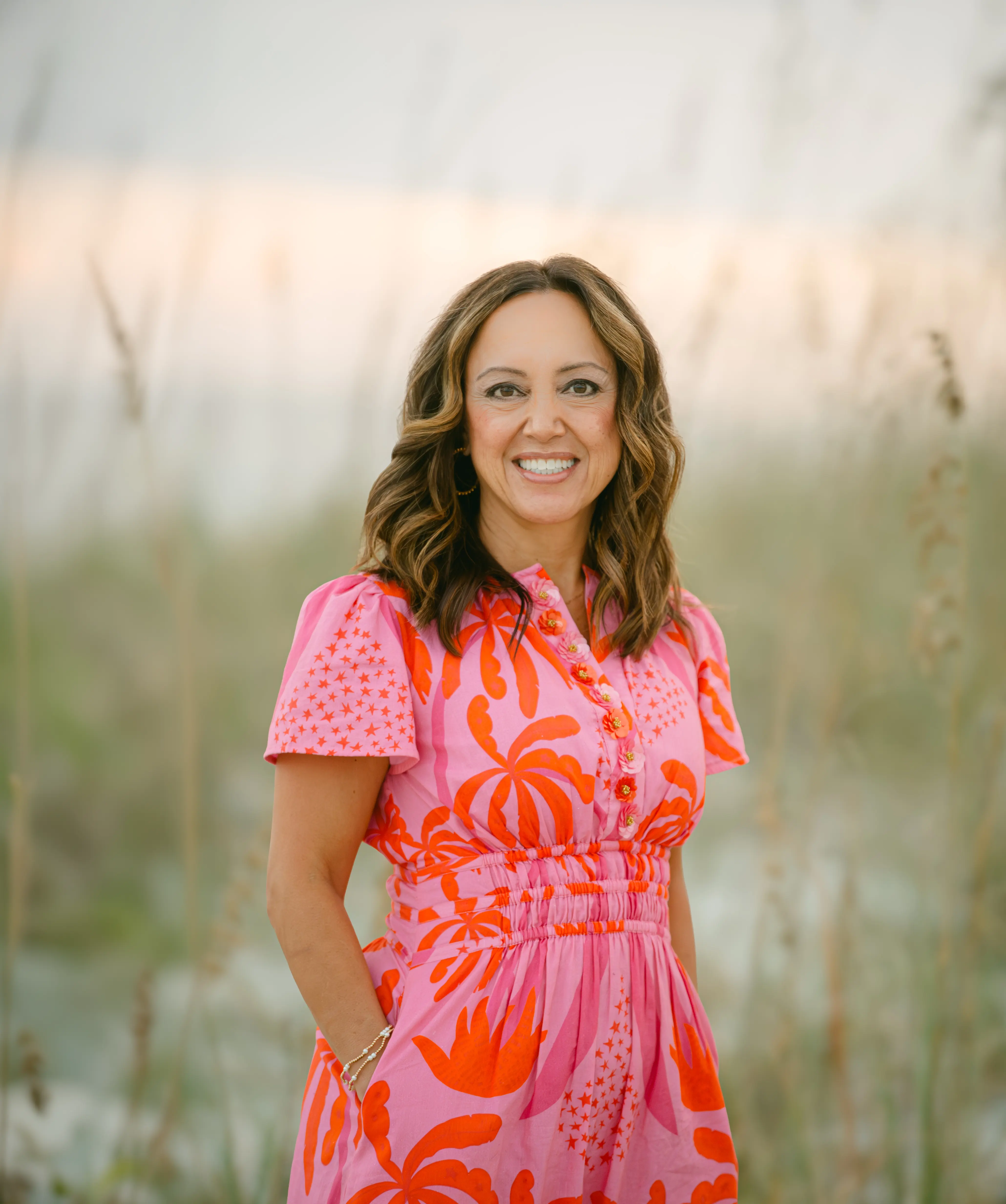 Woman in a pink and orange floral dress standing in coastal dune grass at sunset