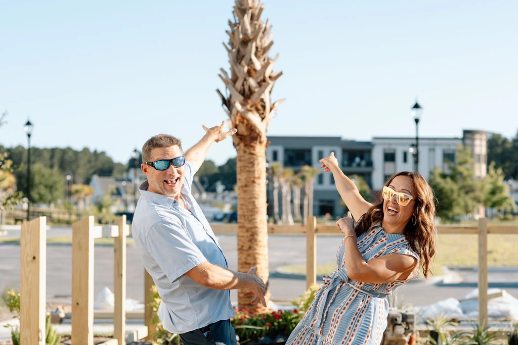 A man and woman celebrating outdoors with arms raised near a palm tree