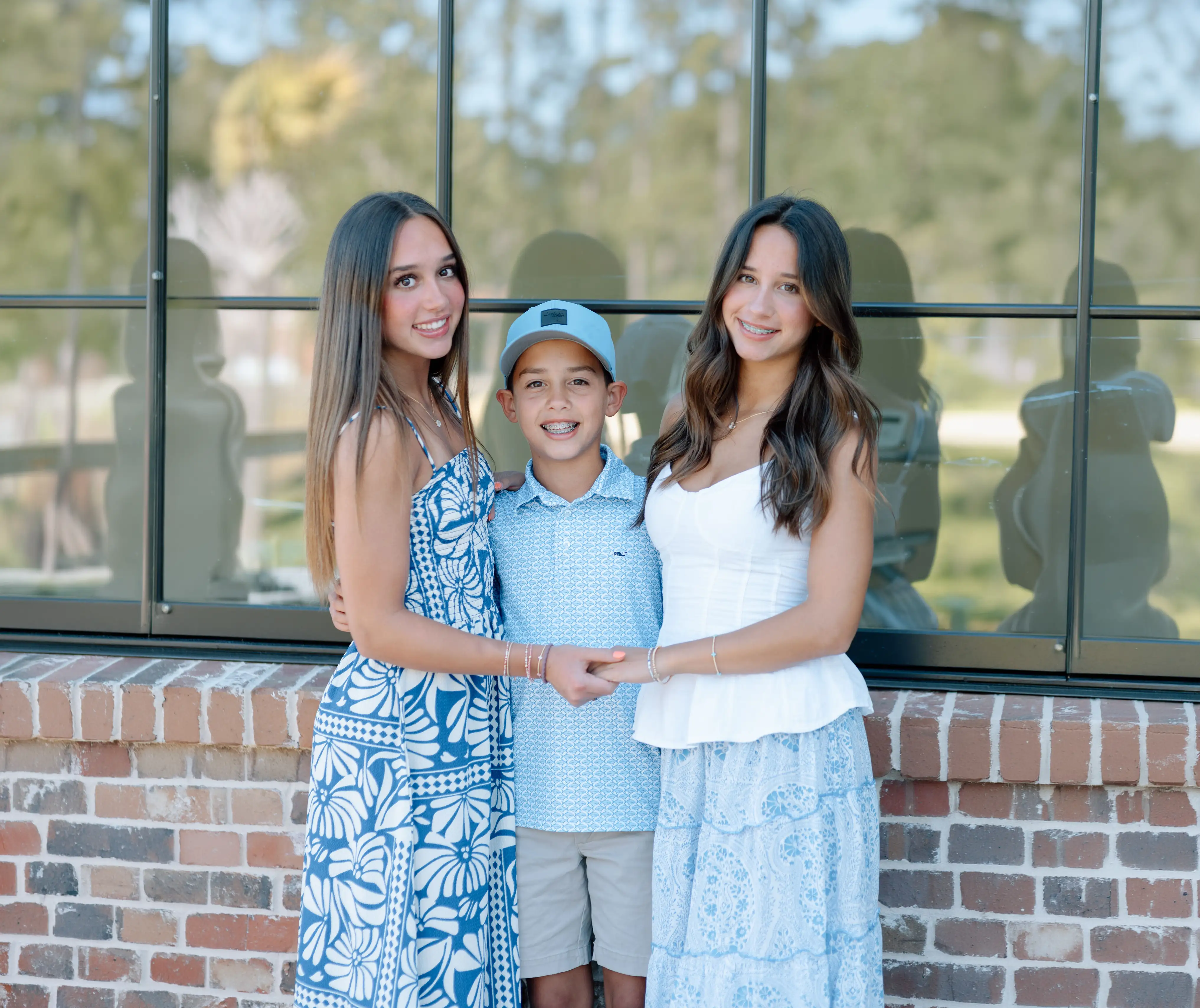 Two teenage girls and a young boy standing together outdoors near a glass-walled building