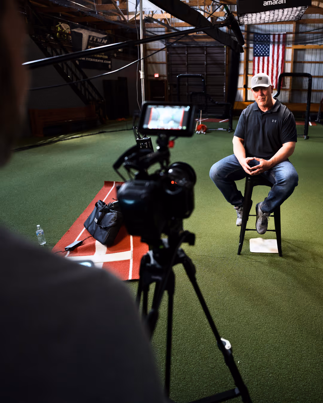 Man in a baseball cap sitting on a stool on turf, filmed by a camera in an indoor sports facility.