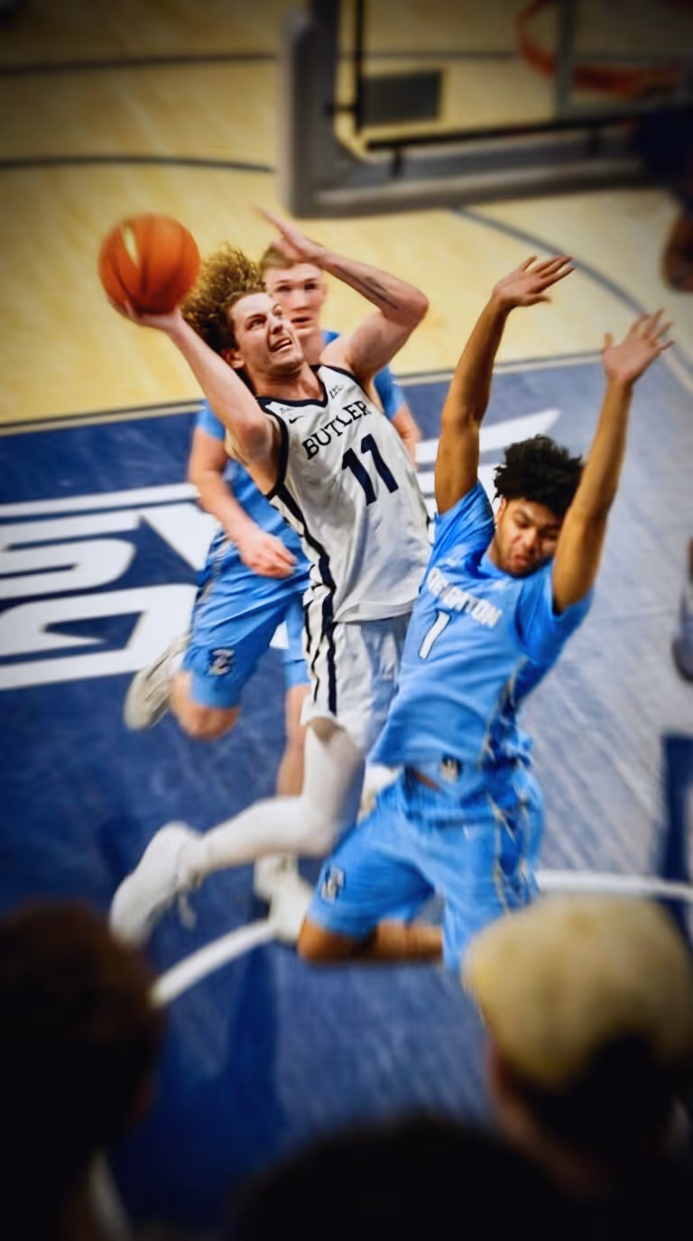 Butler basketball player in white jersey jumps to shoot while two Creighton defenders block him.