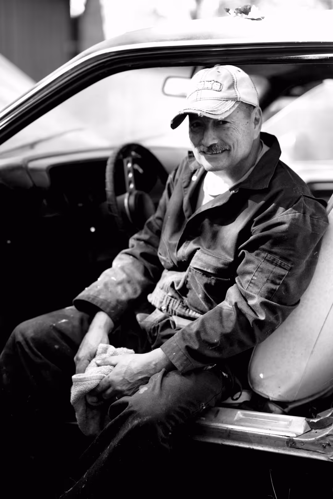 Smiling man in a cap and coveralls sitting in a car holding a cloth, black and white photo.