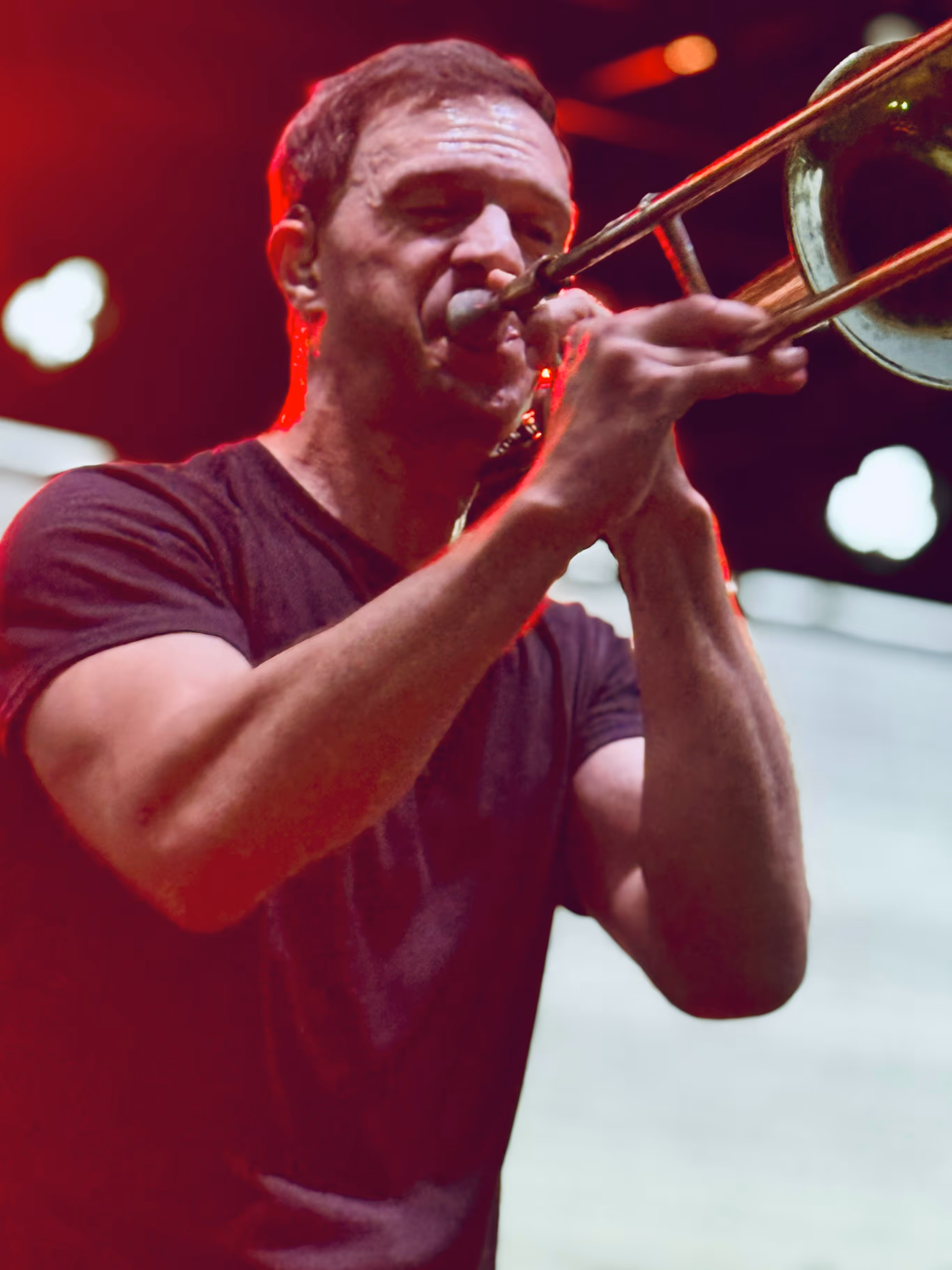 Man in black t-shirt playing a trombone with stage lights in the background.