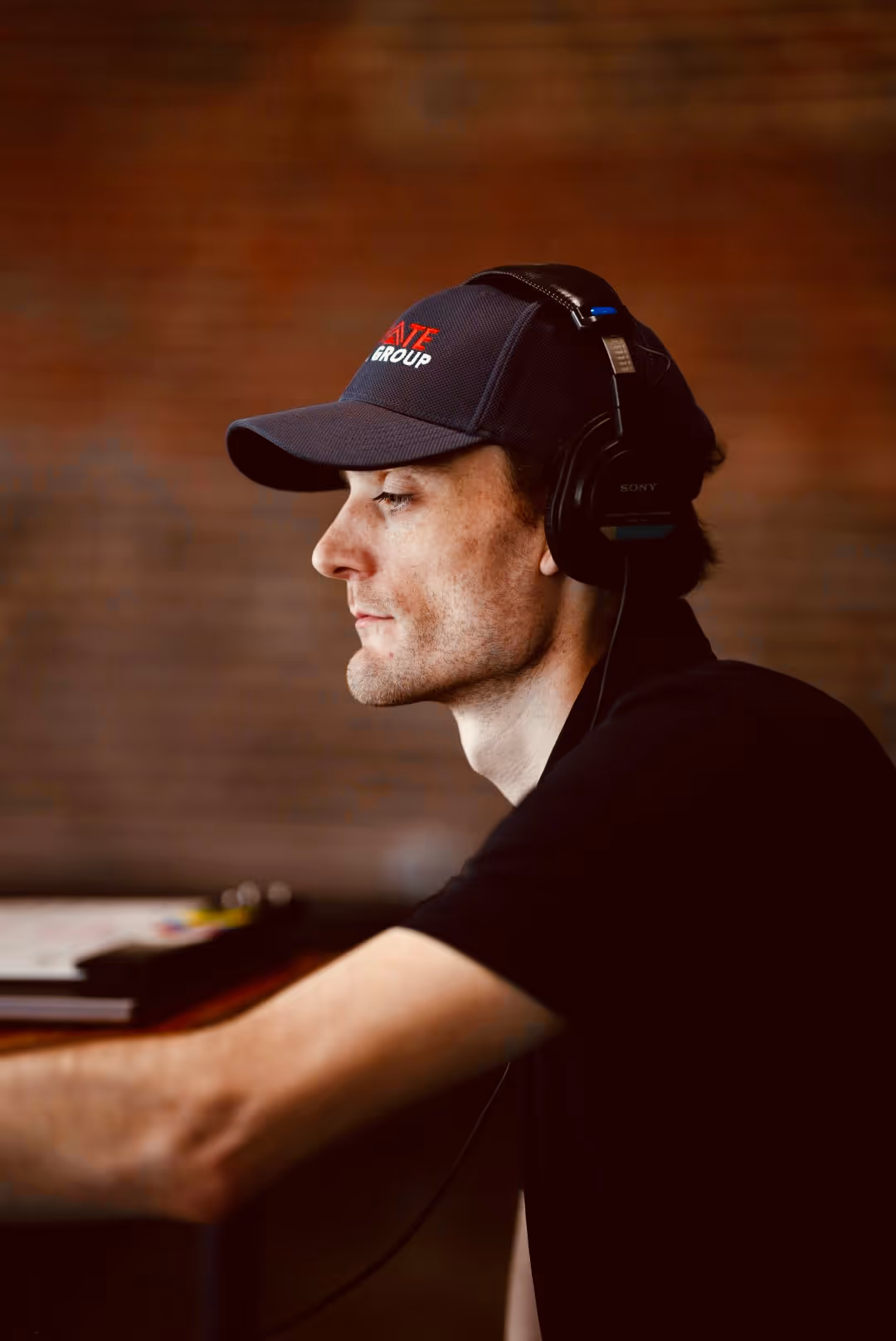 Side profile of a young man wearing headphones and a dark cap with red and white text indoors.