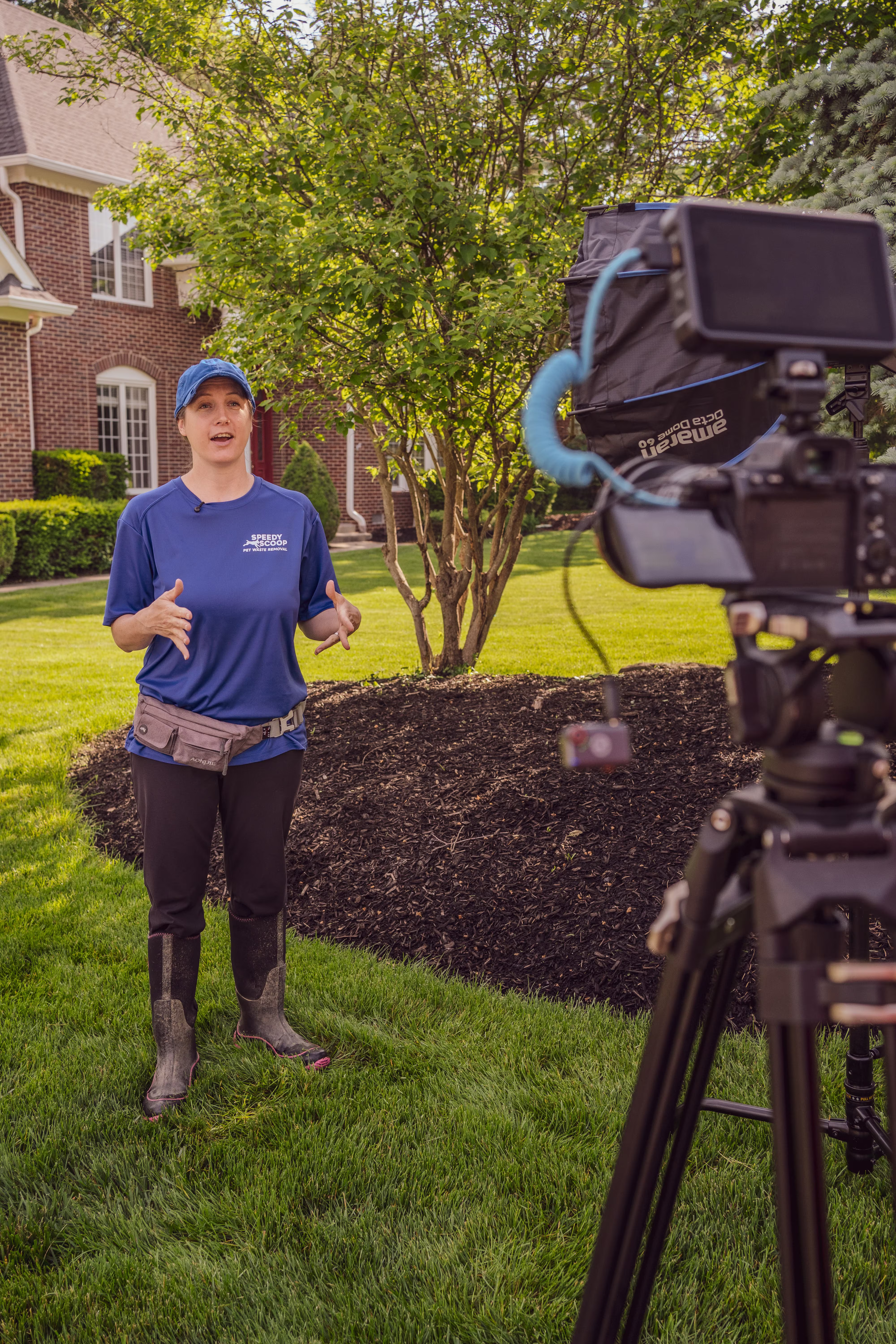 Woman in blue shirt and cap talking on lawn in front of video camera with lighting equipment.