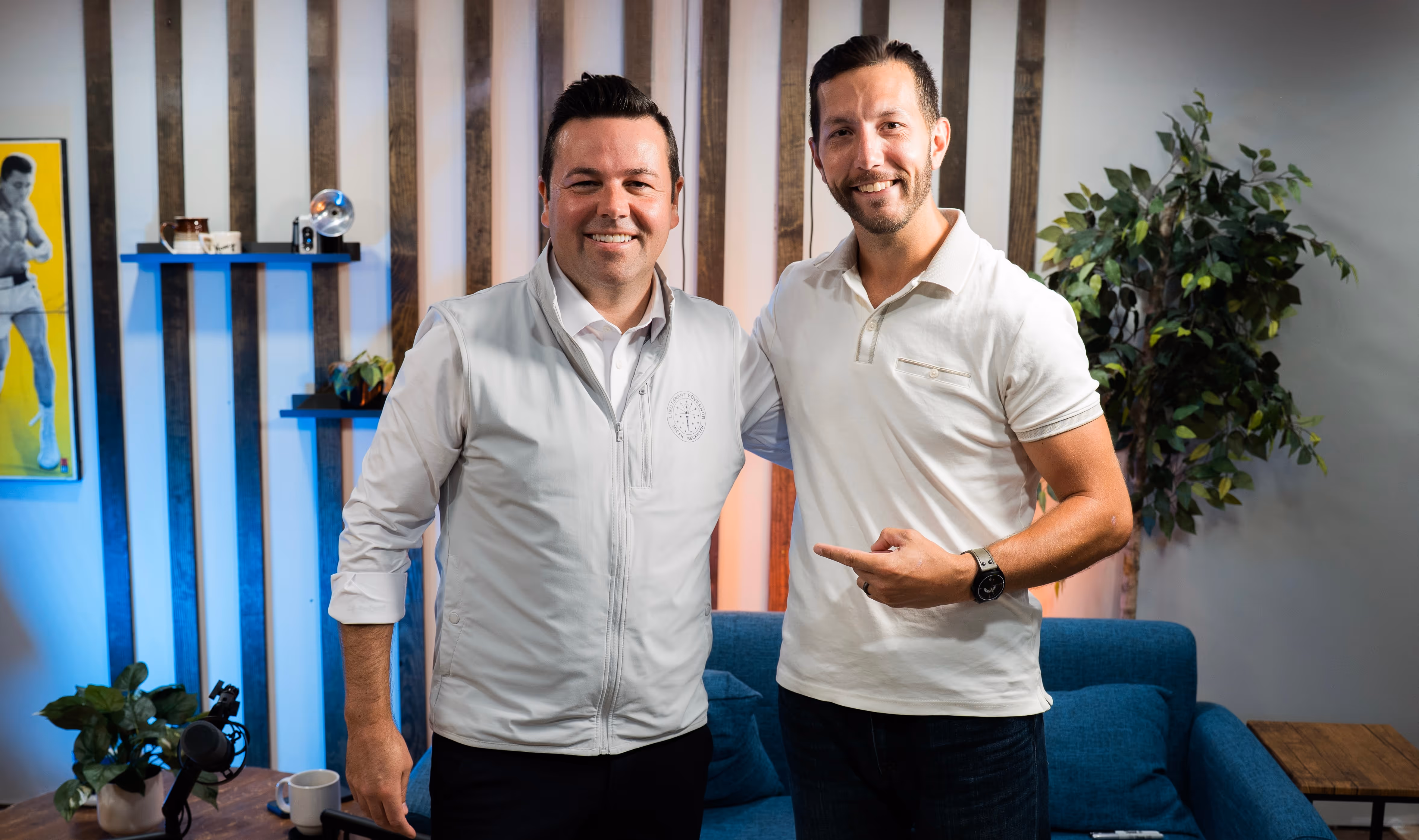Two smiling men standing in a modern living room with plants, artwork, and blue couch.