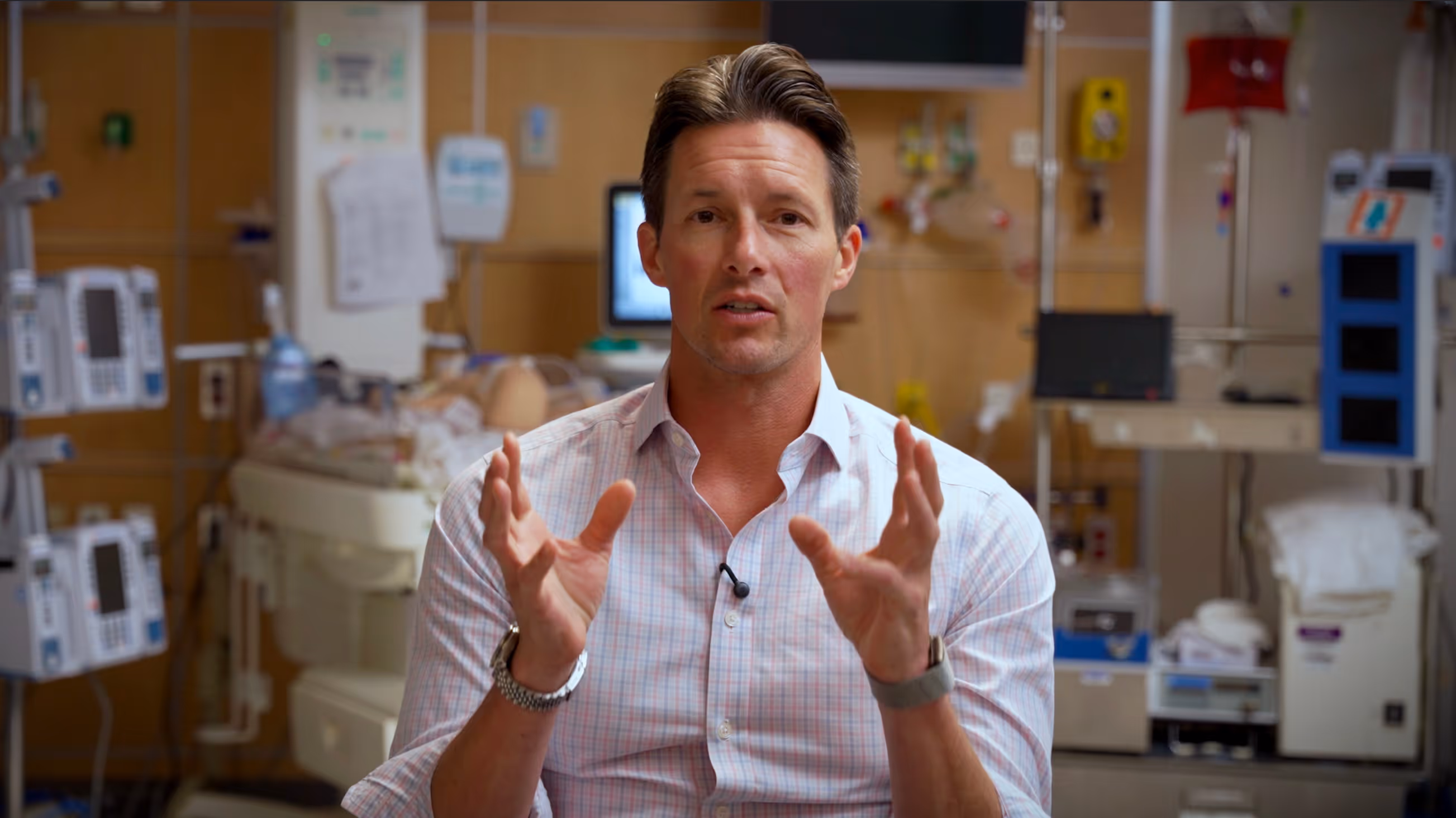 Man in a checkered shirt speaking with hands raised in a hospital room with medical equipment.