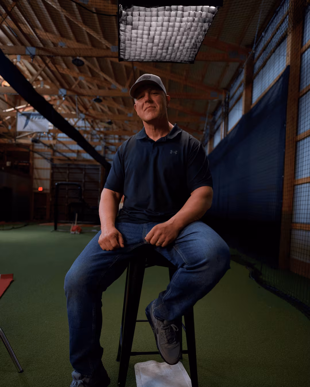 Man wearing a cap, dark shirt, and jeans sitting on a stool in an indoor sports facility.