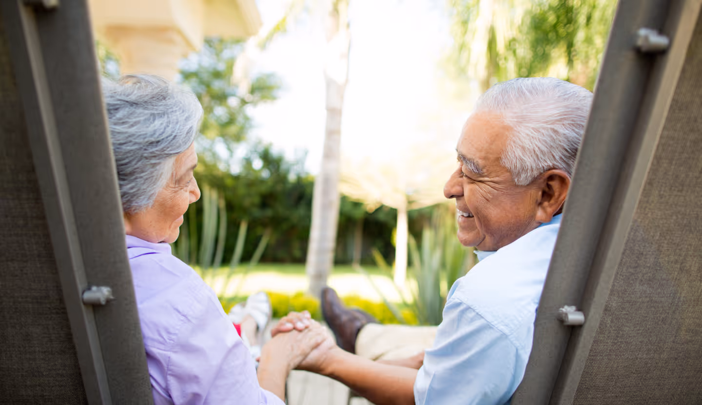 Senior couple holding hands and sitting outdoors on lounge chairs in a garden.