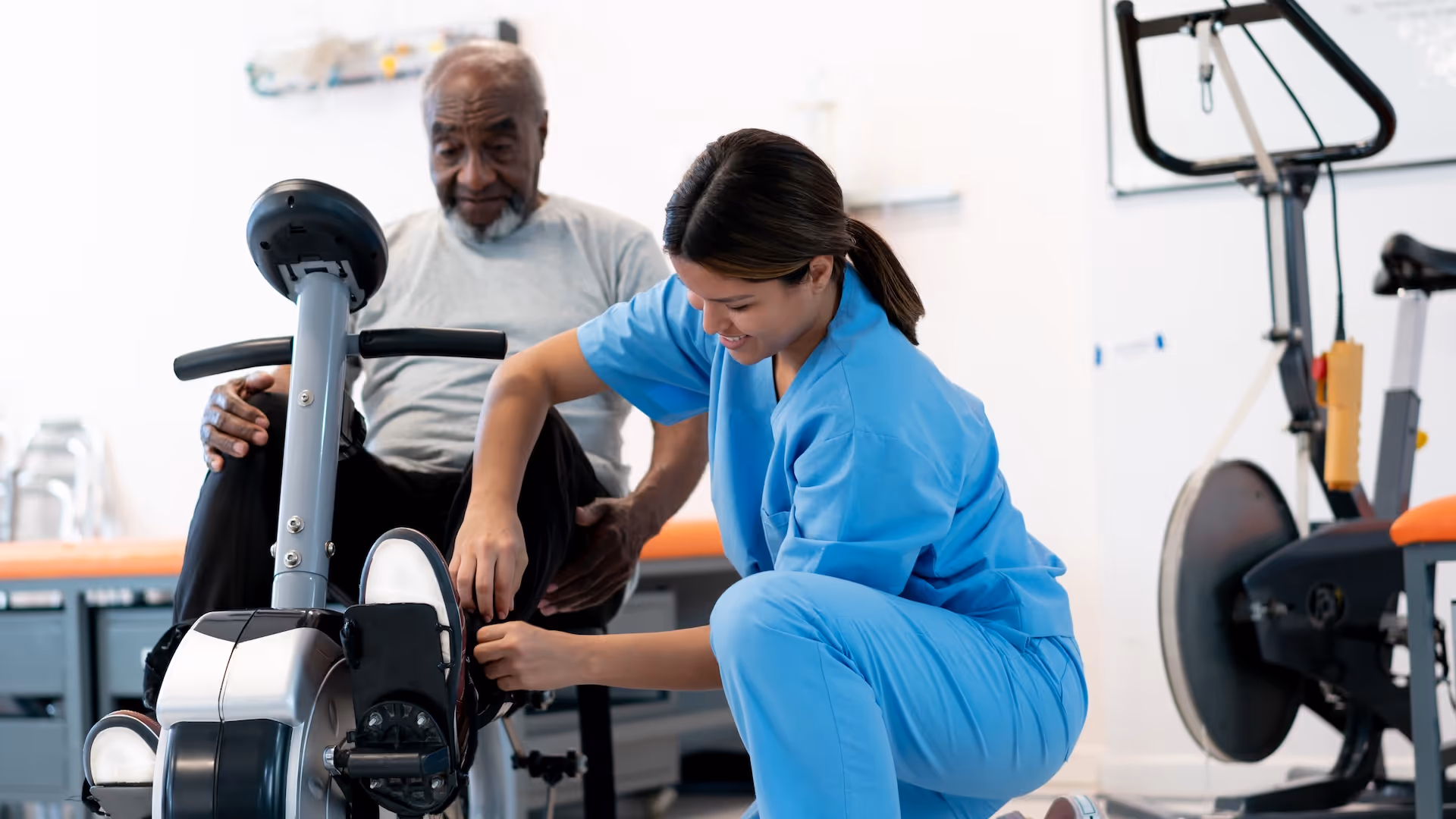 A healthcare worker in blue scrubs adjusting the foot strap of an exercise bike for an elderly man during rehabilitation.