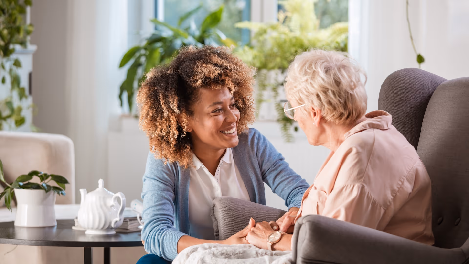 Young woman smiling and holding hands with an elderly woman seated in a chair in a bright room with plants.
