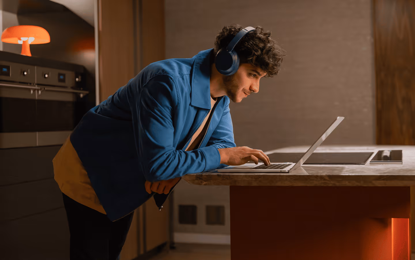 Man wearing blue jacket and headphones leaning on a kitchen counter while using a laptop.