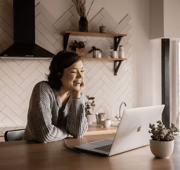 Woman wearing headphones smiling and looking at a laptop while sitting at a wooden kitchen table.
