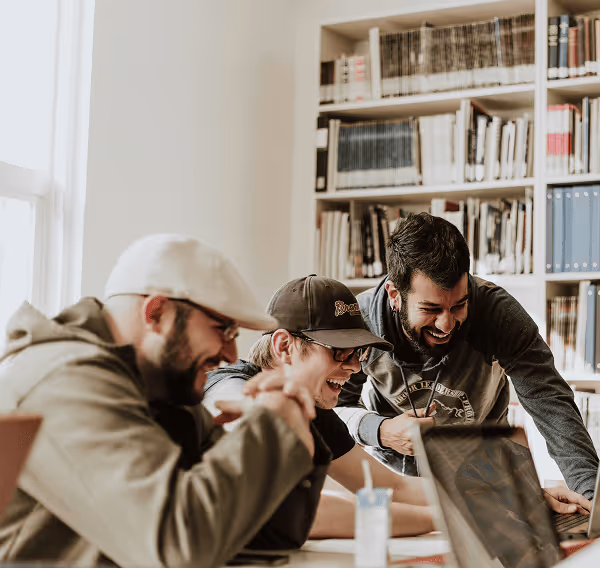 Three young men laughing and looking at a laptop together in a room with bookshelves.