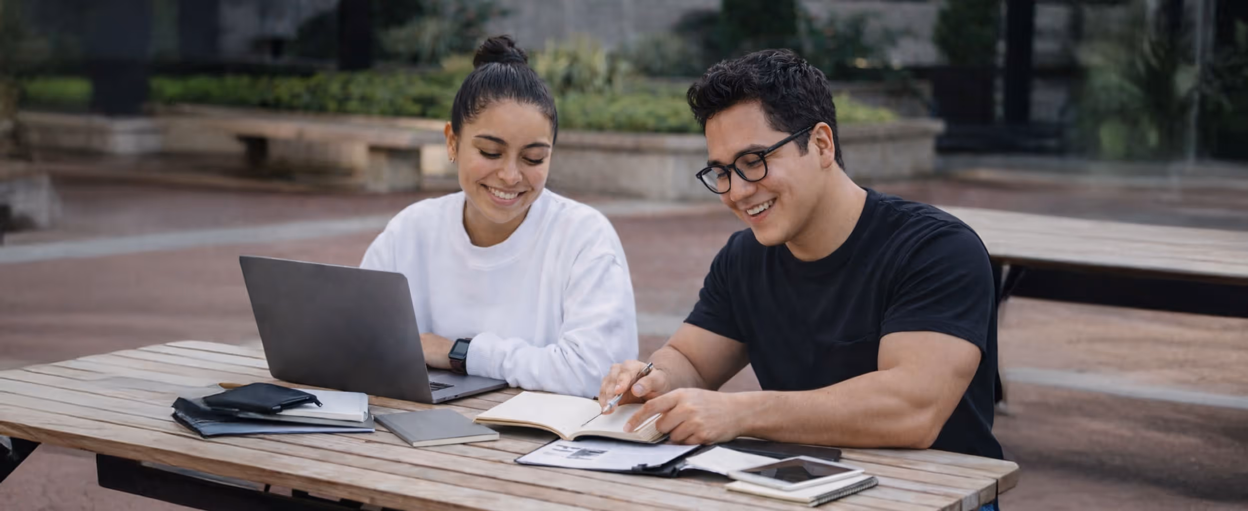 Two young adults sitting at an outdoor wooden table, smiling and studying with a laptop, notebooks, and a smartphone.