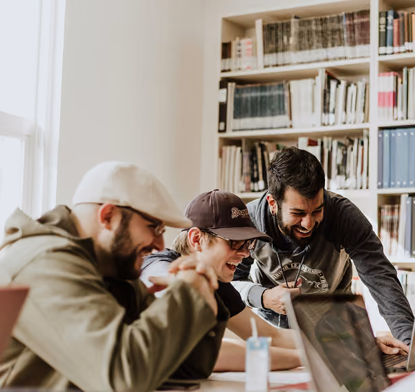 Three young men laughing and working together on laptops in a bright room with bookshelves.