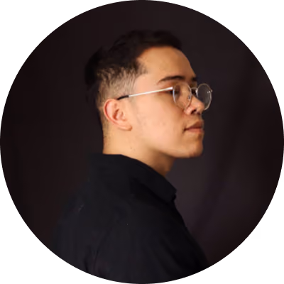Portrait of a young person with short dark hair and round glasses, wearing a black shirt, shown in profile against a dark background.