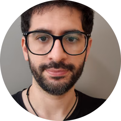 Close-up portrait of a man with short black hair, a beard, and black-framed glasses, wearing a black shirt and necklace, against a plain light background.