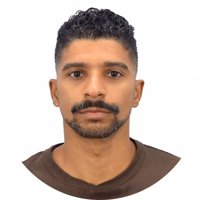 Portrait of a man with short curly black hair, a mustache and goatee, wearing a brown shirt, facing forward with a neutral expression on a white background.