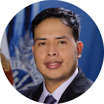 Man with short, dark hair and a light beard, wearing a dark suit, white shirt, and dotted tie, with a blue flag and an emblem in the blurred background.