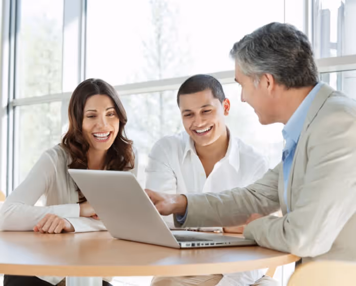 Three people smiling and looking at a laptop on a round table in a bright office.