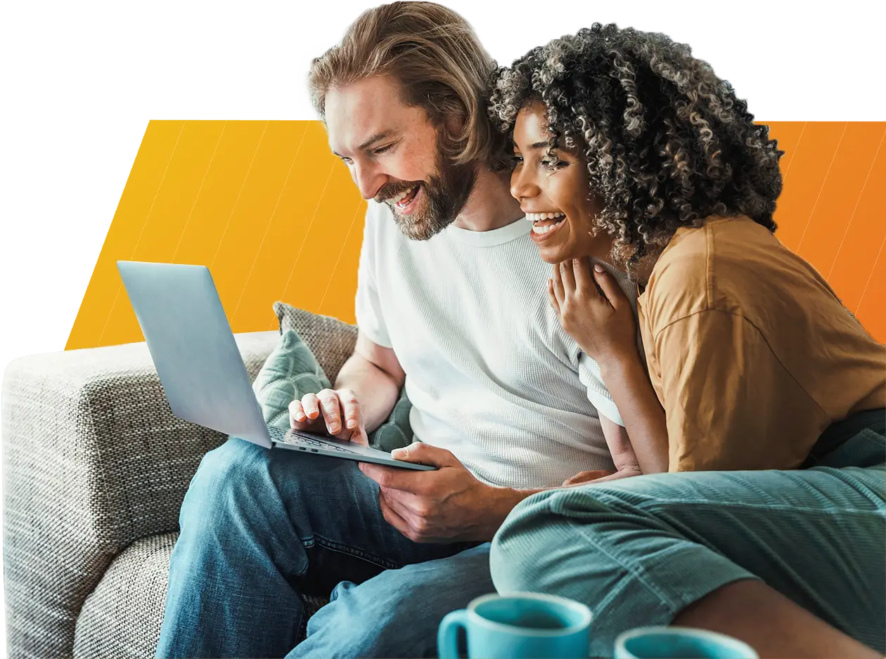 Smiling couple sitting on a couch looking at a laptop together with coffee cups nearby.