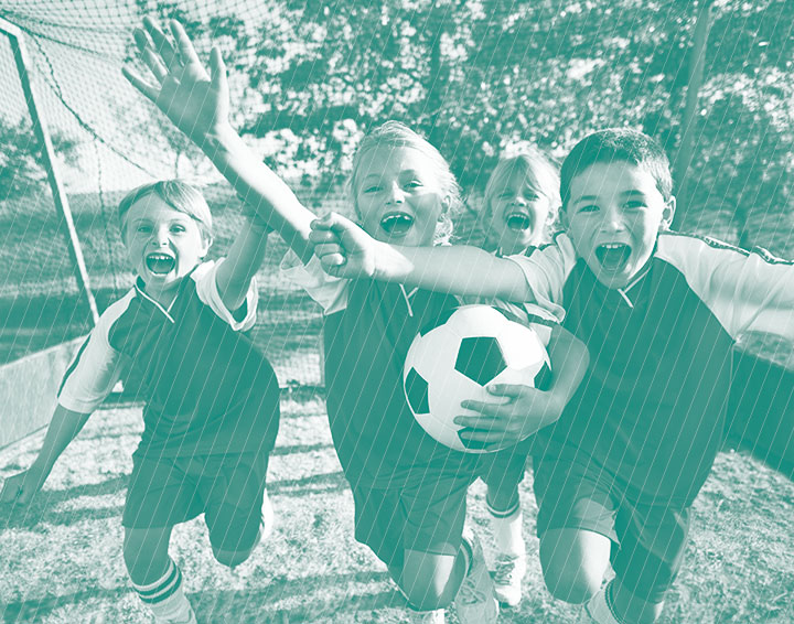 Four excited children in soccer uniforms celebrating near the goal with one holding a soccer ball.
