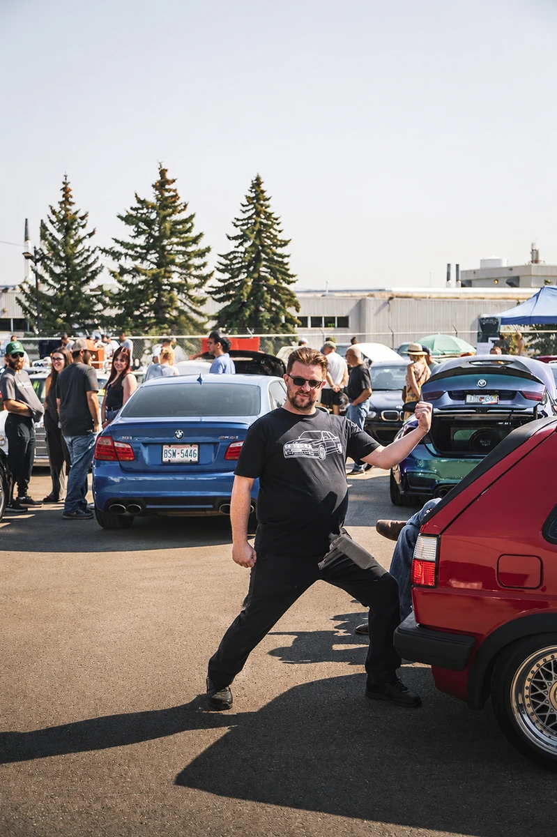 Man striking a playful pose near parked cars at an outdoor car event with pine trees in the background.
