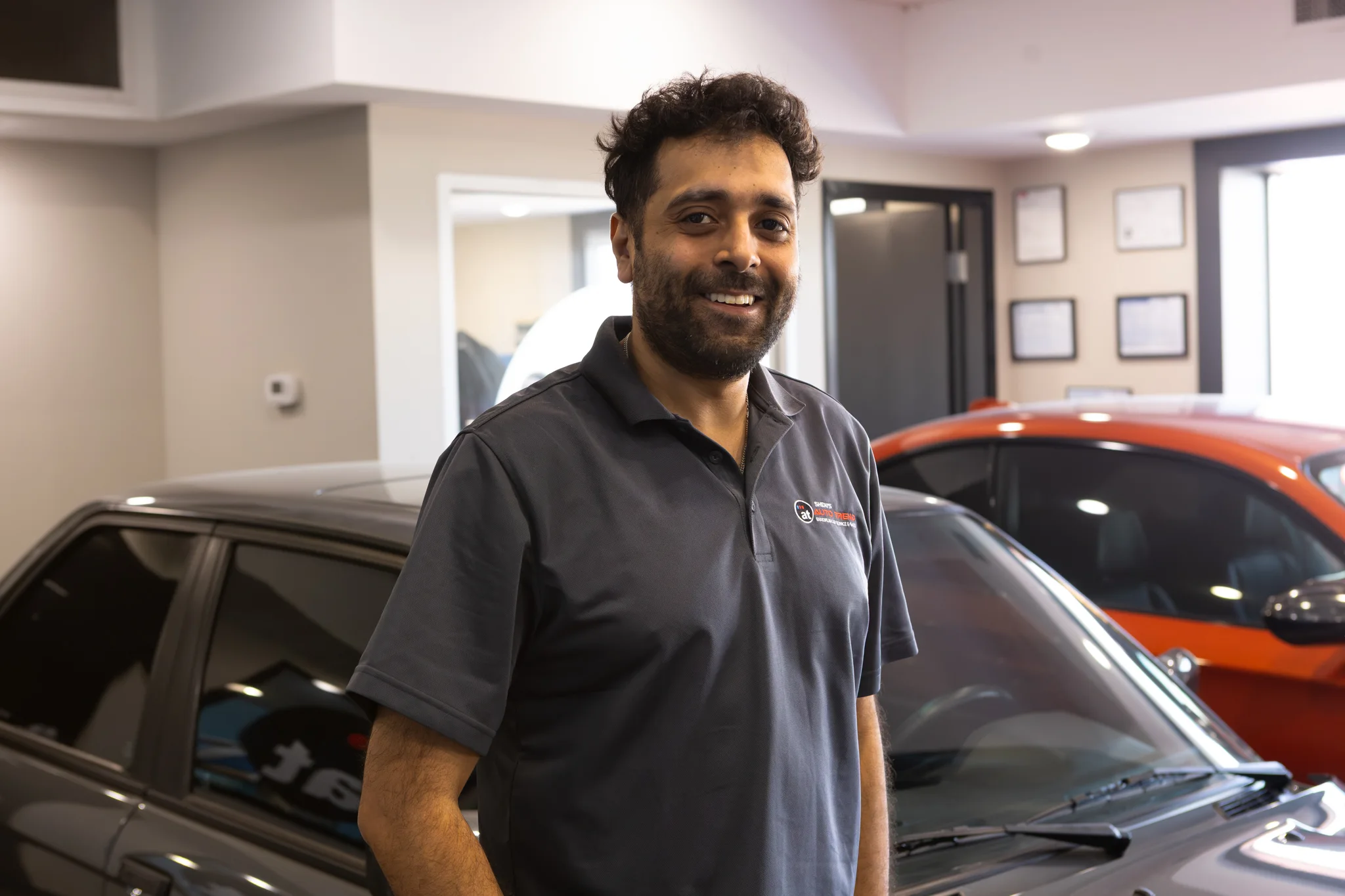 Man in a polo shirt smiling inside a garage with a black and an orange car behind him.