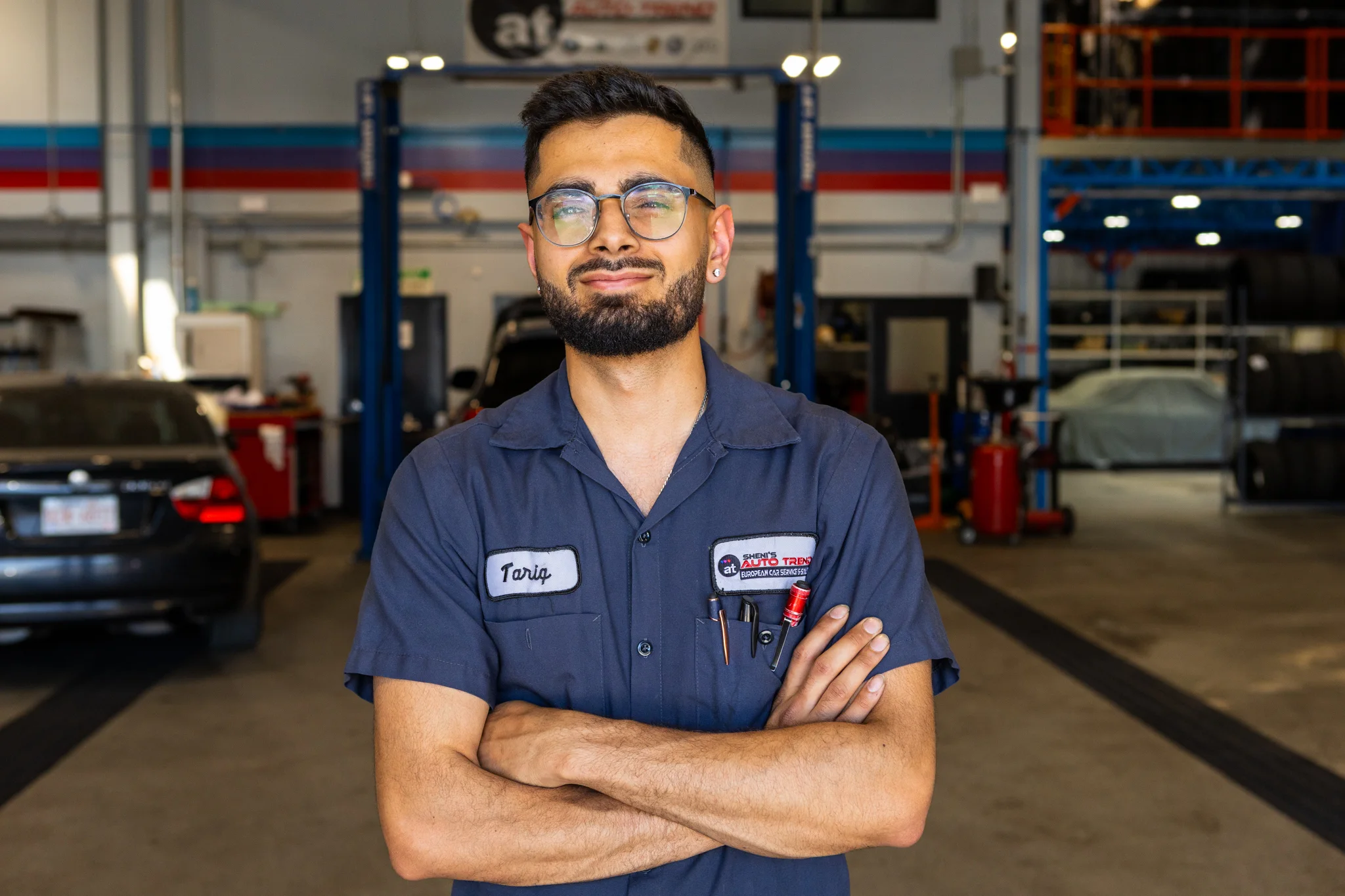 Mechanic with glasses and beard standing with arms crossed in an auto repair shop.