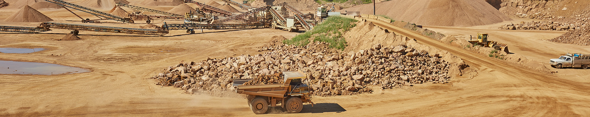 Quarry site with heavy machinery, large piles of rocks and gravel, and a truck in motion on dirt ground.