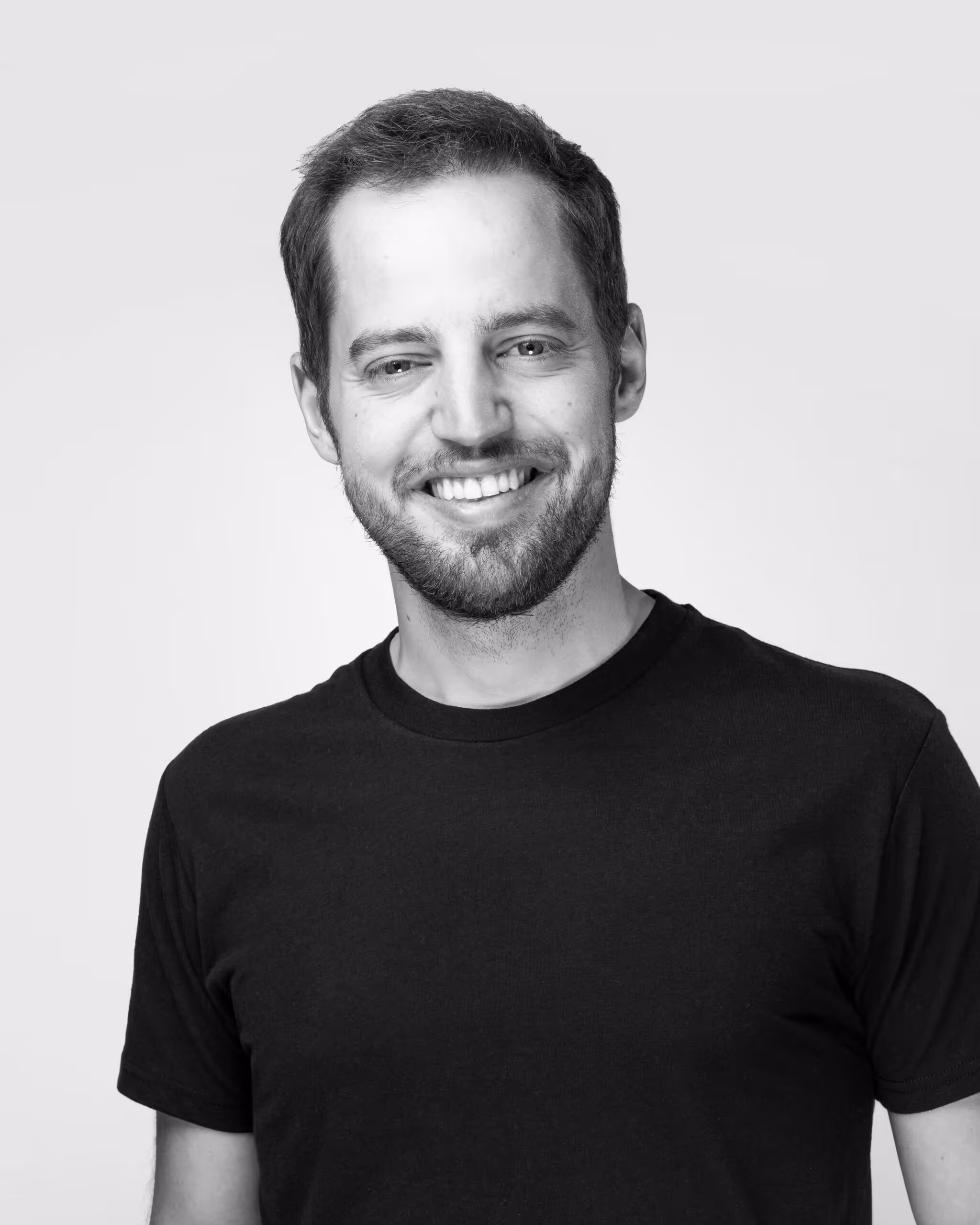 Smiling man with short brown hair, beard, and blue eyes wearing a black shirt against a black background.