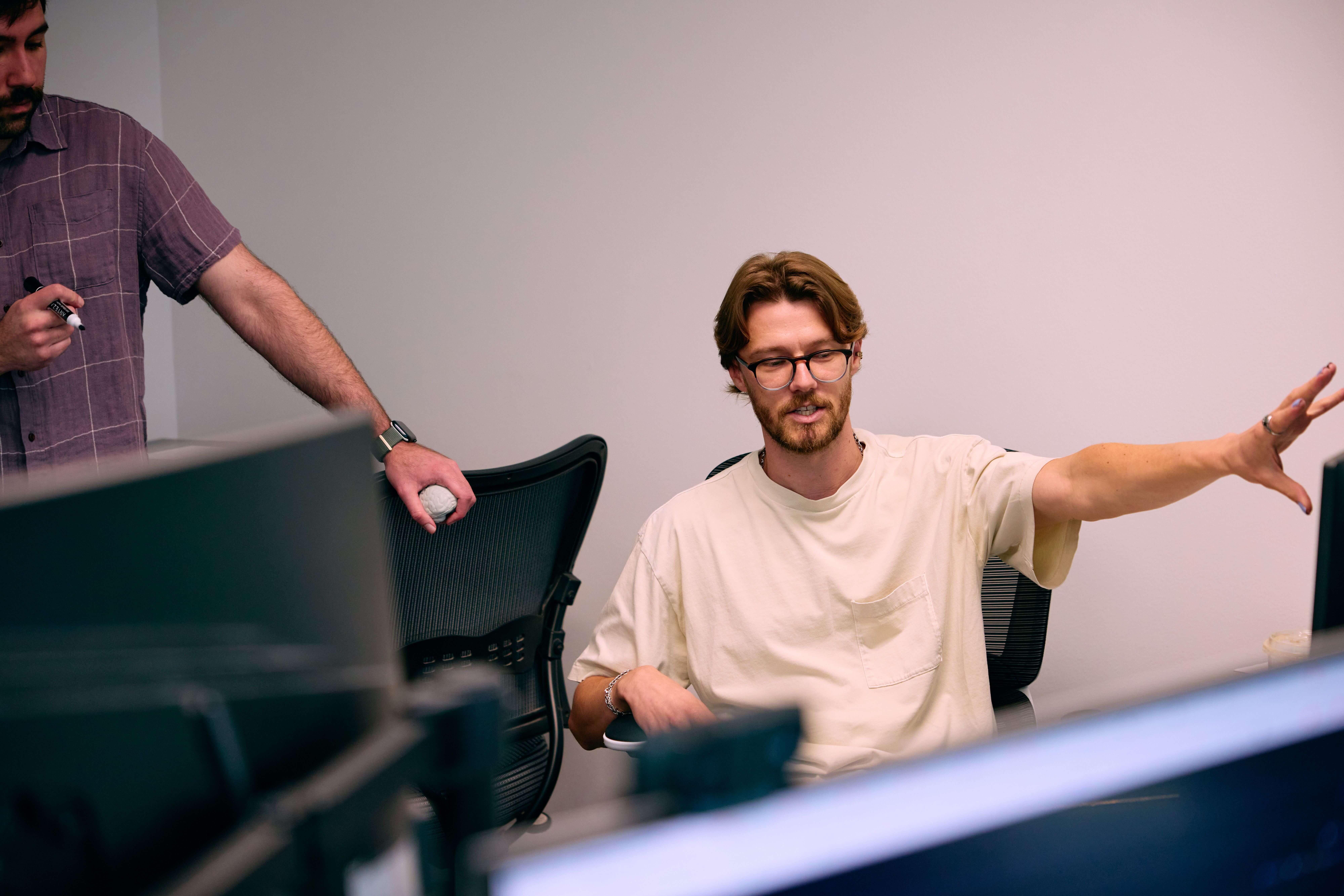 Man with glasses in a white t-shirt gesturing with his hand while another man in a checked shirt holds a marker, both in an office setting.