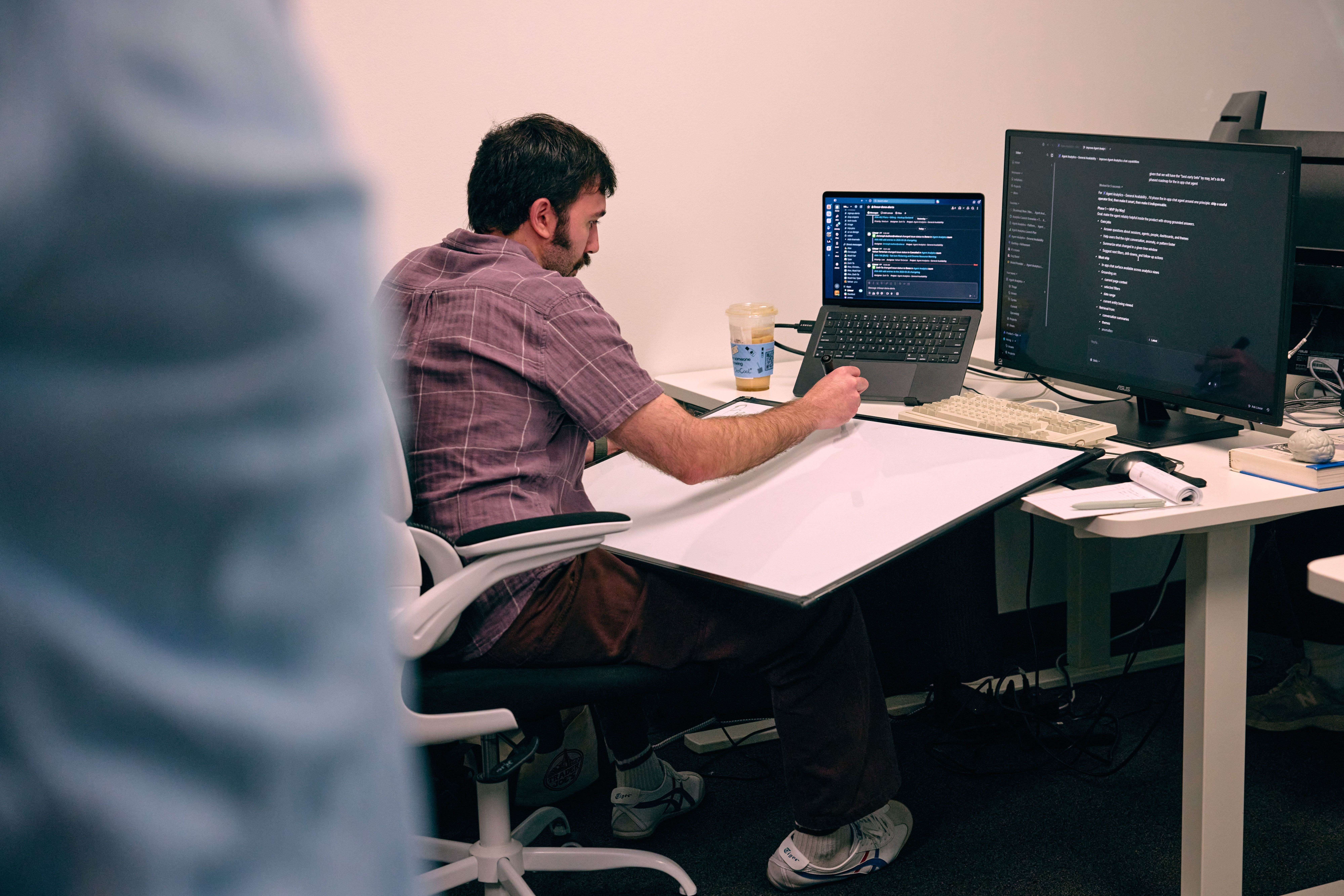 Man sitting at desk working on a laptop and a desktop monitor displaying code and a messaging app.