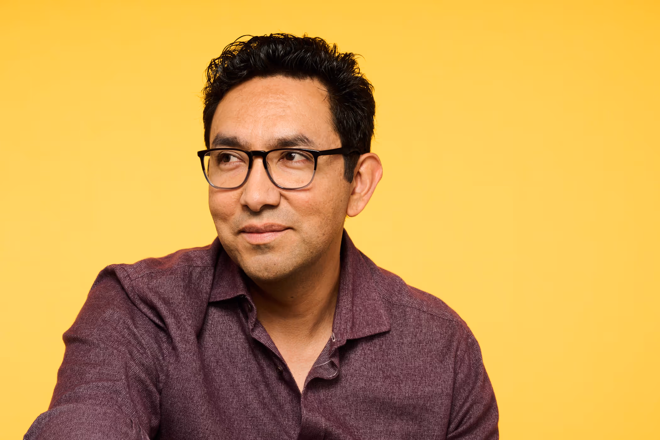Man with black glasses and dark hair wearing a maroon shirt looking to the side against a yellow background.