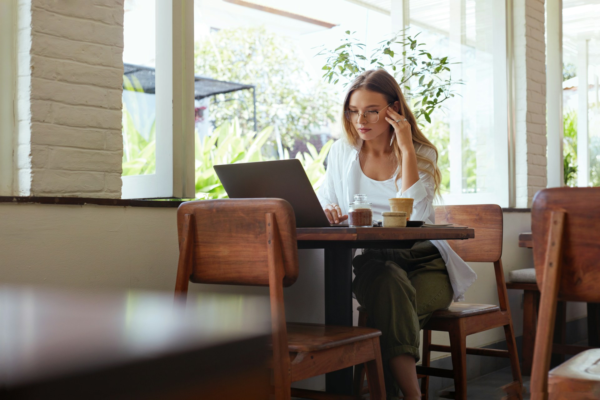 Woman wearing glasses working on laptop at a wooden table in a bright cafe near a window.