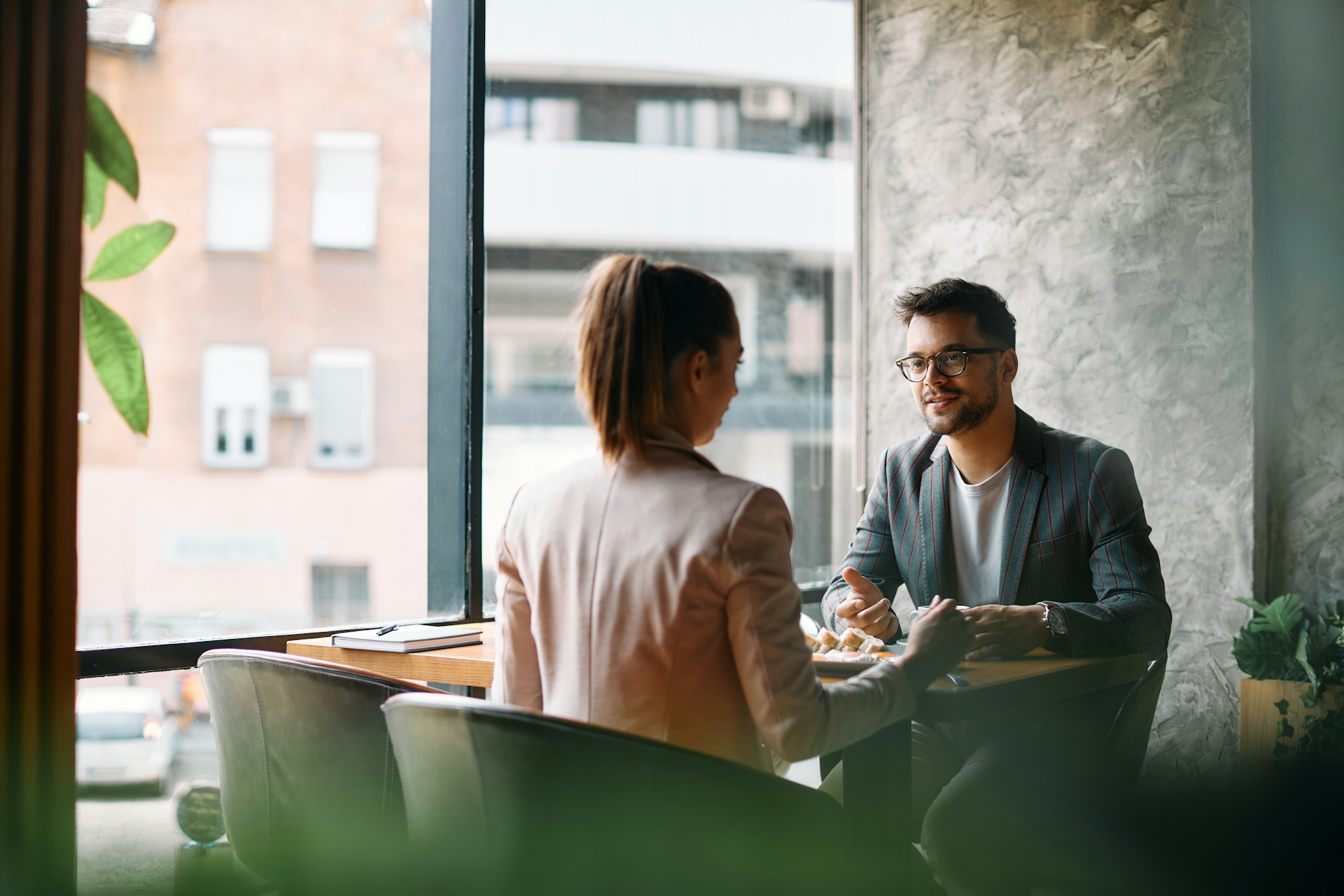 Man and woman talking and eating at a table near a large window in a modern cafe.