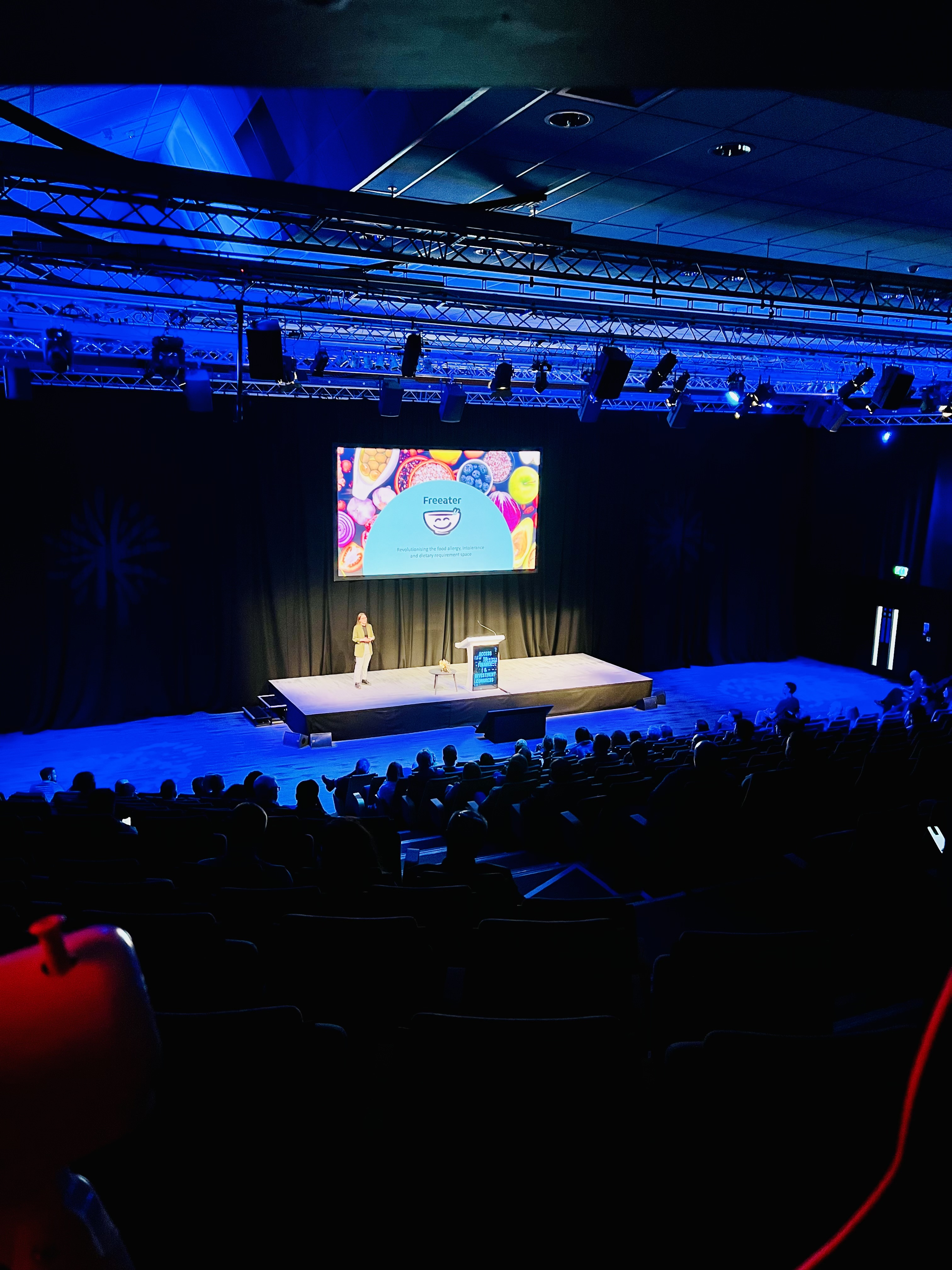 Momentum Valley Finals Pitch. Kate the founder pitching on stage with Freeater presentation in the background. View of the audience from a far.