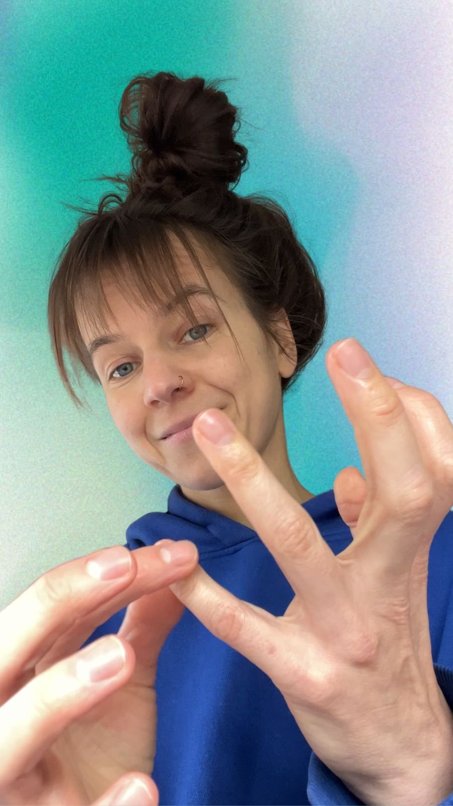 Woman with nose ring and hair in a messy bun wearing a blue hoodie, showing her hands in a close-up shot against a teal and light purple gradient background.
